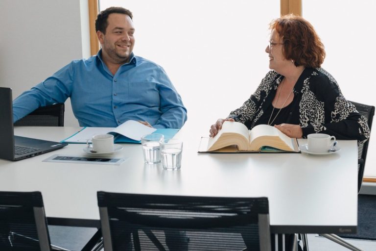 Steuerberatung Frau Pestel und Herr Pestel sitzen an einem Tisch und unterhalten sich über die Steuerberatung, Bücher und Laptop sind vorhanden.