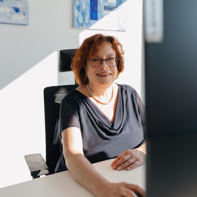 Sandra Pestel mit Brille sitzt am Schreibtisch und lächelt in die Kamera.