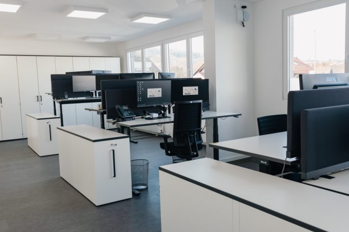 Moderne Büroräume mit Schreibtischen und Computerbildschirmen. Helle Fenster sorgen für Licht.