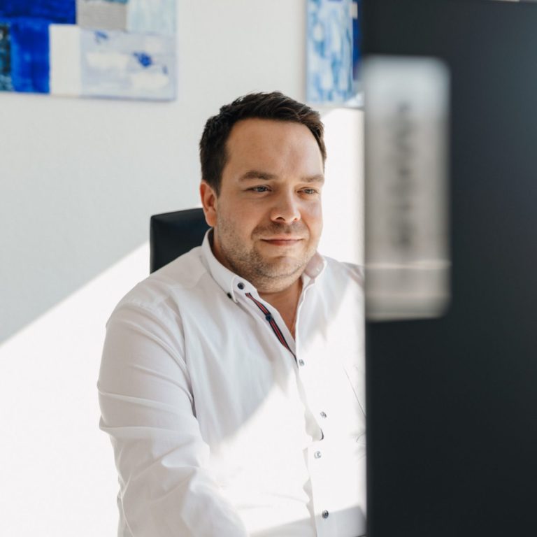 Norman Pestel in weißem Hemd sitzt an einem Schreibtisch und schaut auf einen Computerbildschirm.